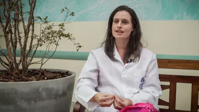 Foto de Karoline Apolônia, uma mulher branca, de cabelos castanhos e lisos, vestida de jaleco sentada em um banco enquanto fala.