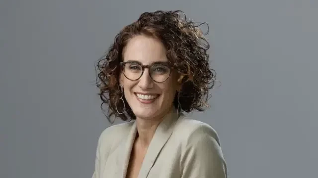 Ana Carolina Evangelista de cabelos cacheados curtos, óculos e terninho bege, sorrindo para a câmera