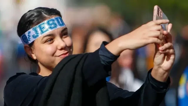 Mulher jovem com uma faixa na testa escrita Jesus tirando selfie com o celular durante a Marcha para Jesus em maio de 2024