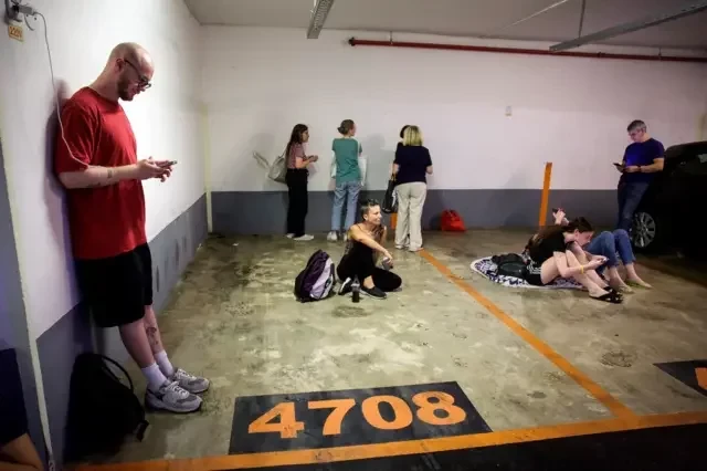 Pessoas em estacionamento, uma sentada, outras conversando, outro carregando o celular