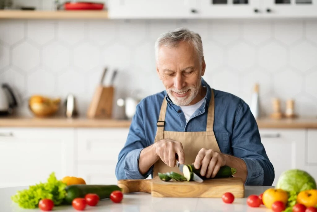 Homem com o cabelo e a barba grisalho usando camisa jeans e avental bege sentado em bancada cortando pepino para salada